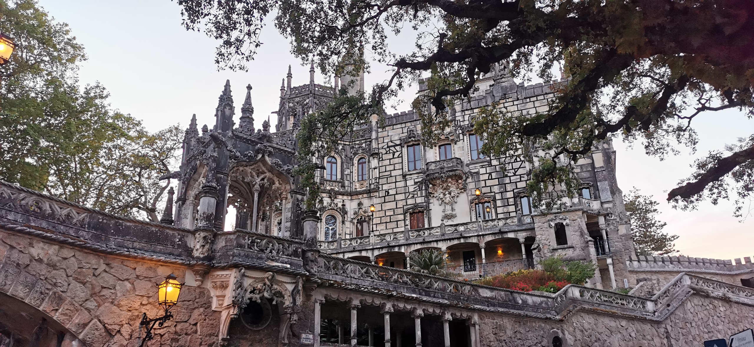 The spired stone facade of Quinta da Regaleira above a mossy retaining wall, with a lit street lamp on the terrace at dusk