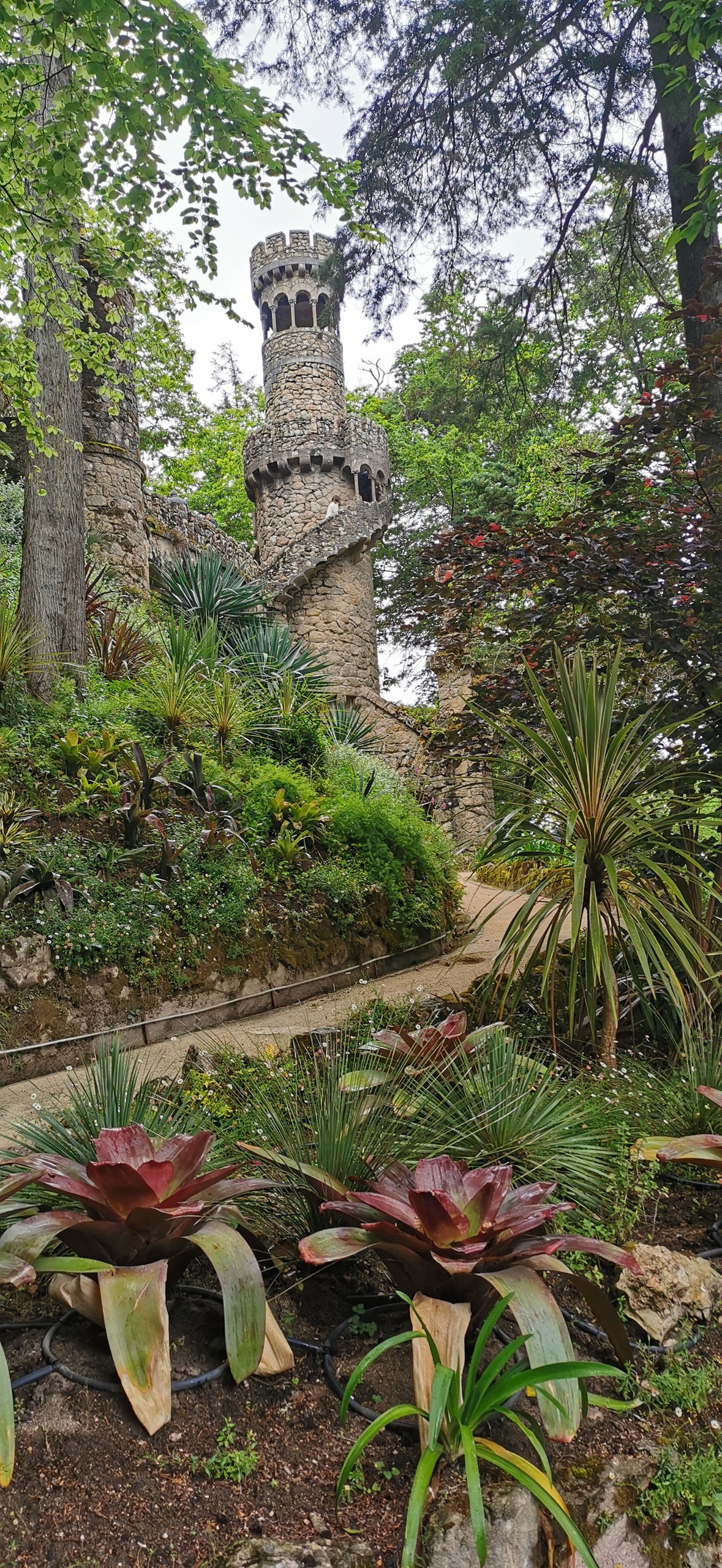 A round stone garden tower at Quinta da Regaleira with an external spiral staircase, set among ferns and a winding dirt path
