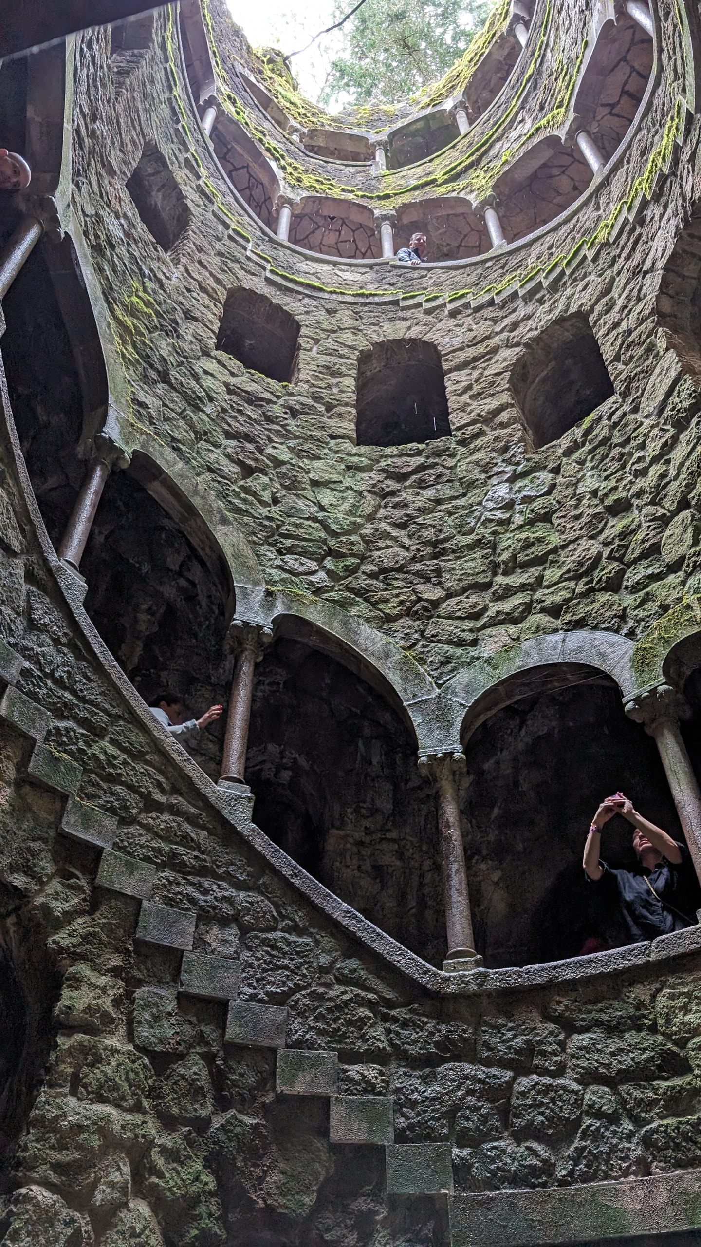Looking up from the floor of the Initiation Well toward a circular opening of sky, with mossy gothic arches and visitors on the spiral stairs