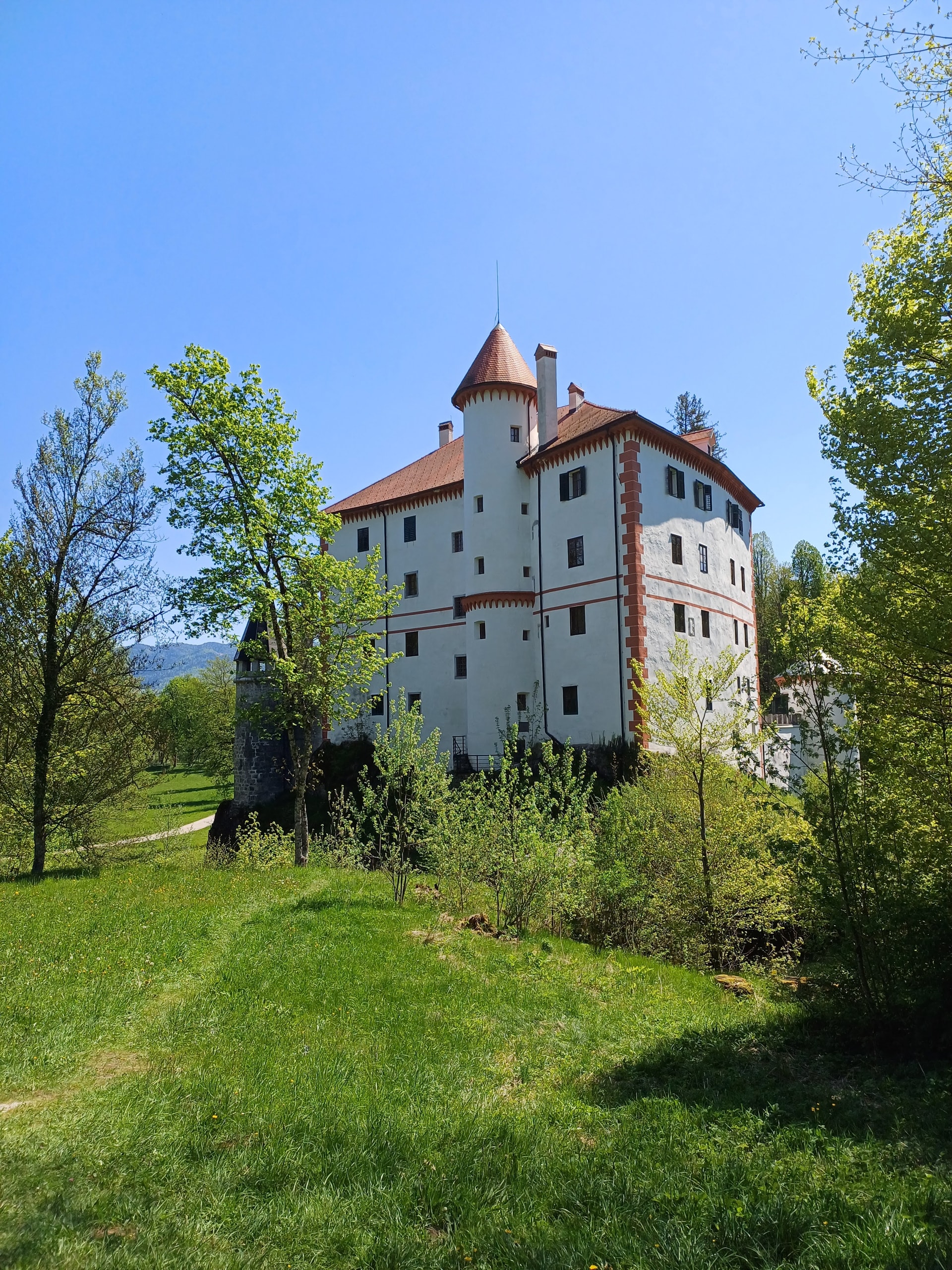 Snežnik Castle and its pointed tower rising above a mown park meadow under a clear blue sky