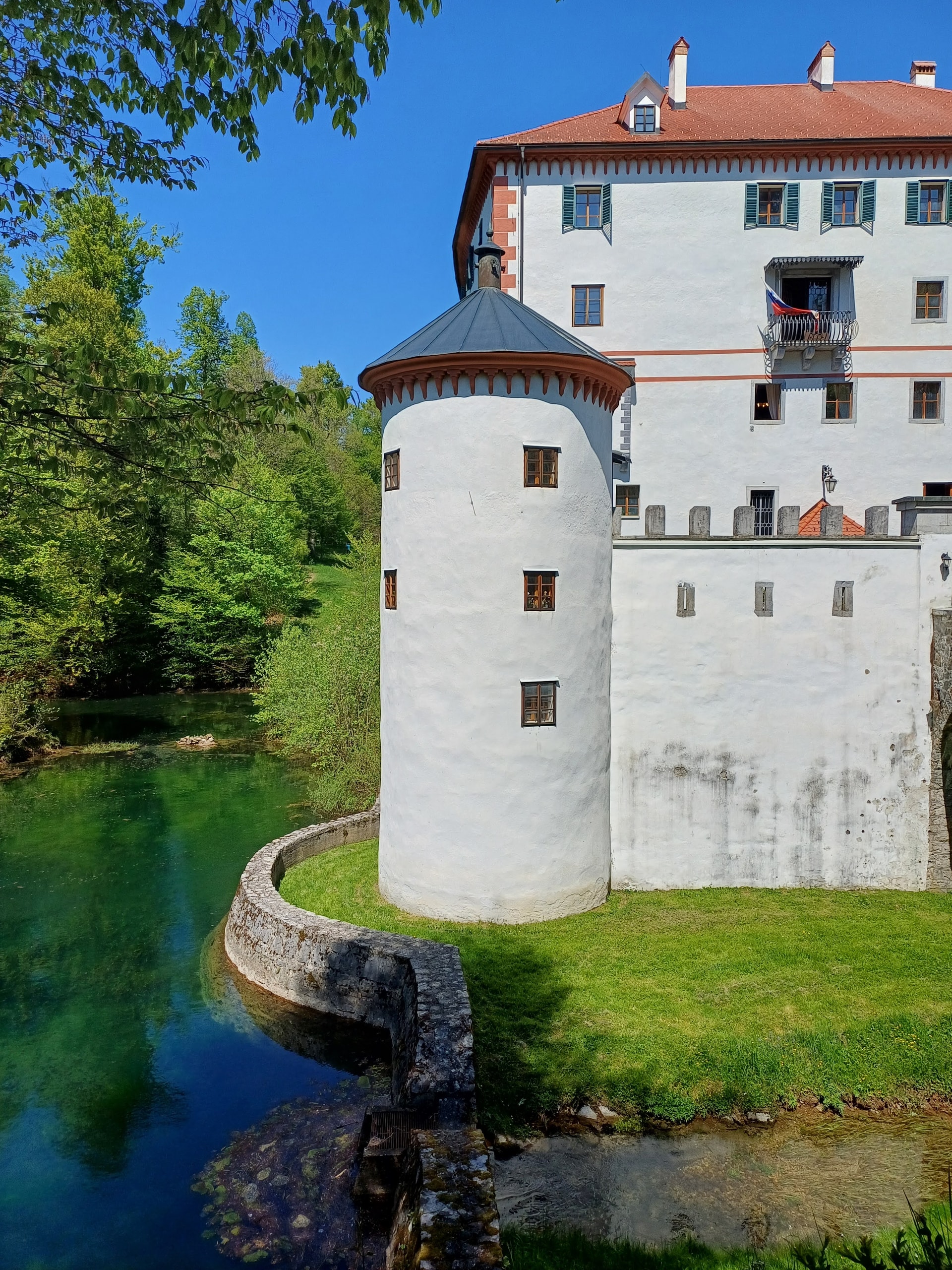 The white castle wall and round tower above a stone arch bridge crossing bright green spring water