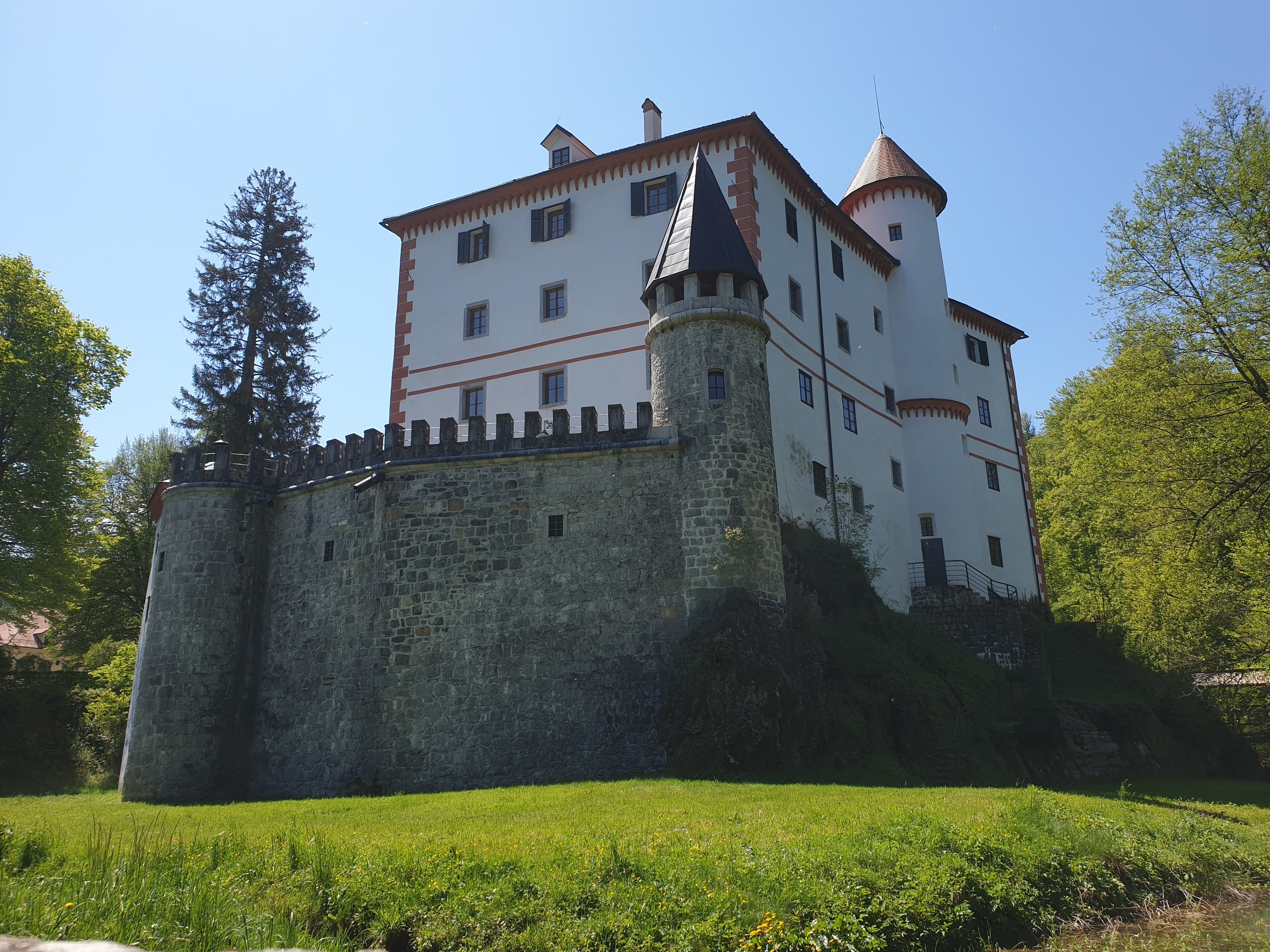 Grey stone curtain wall with square battlements and round towers below the white castle, seen from a spring meadow