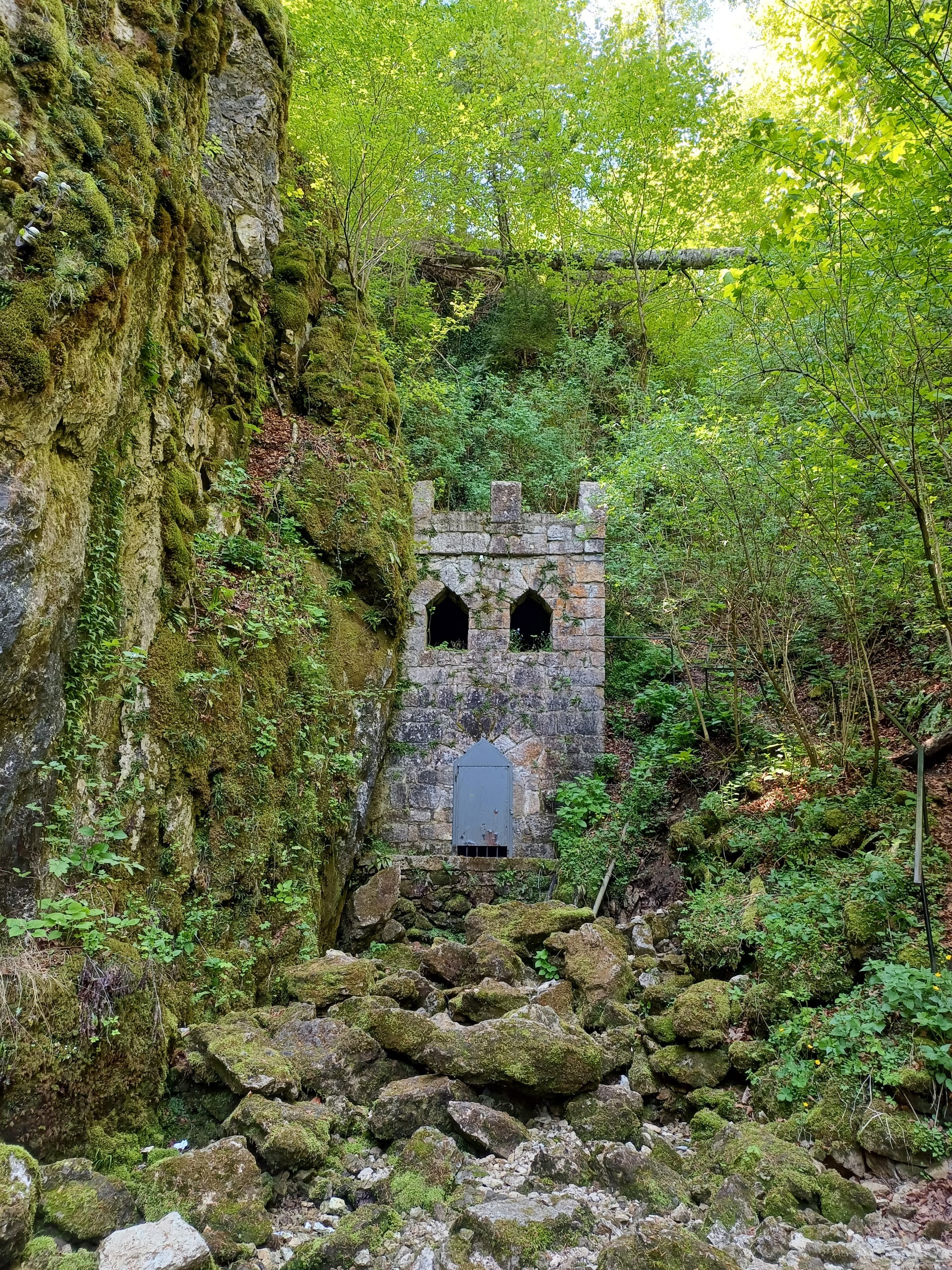 Small stone building with a metal door set into a wall of mossy limestone under bright green trees