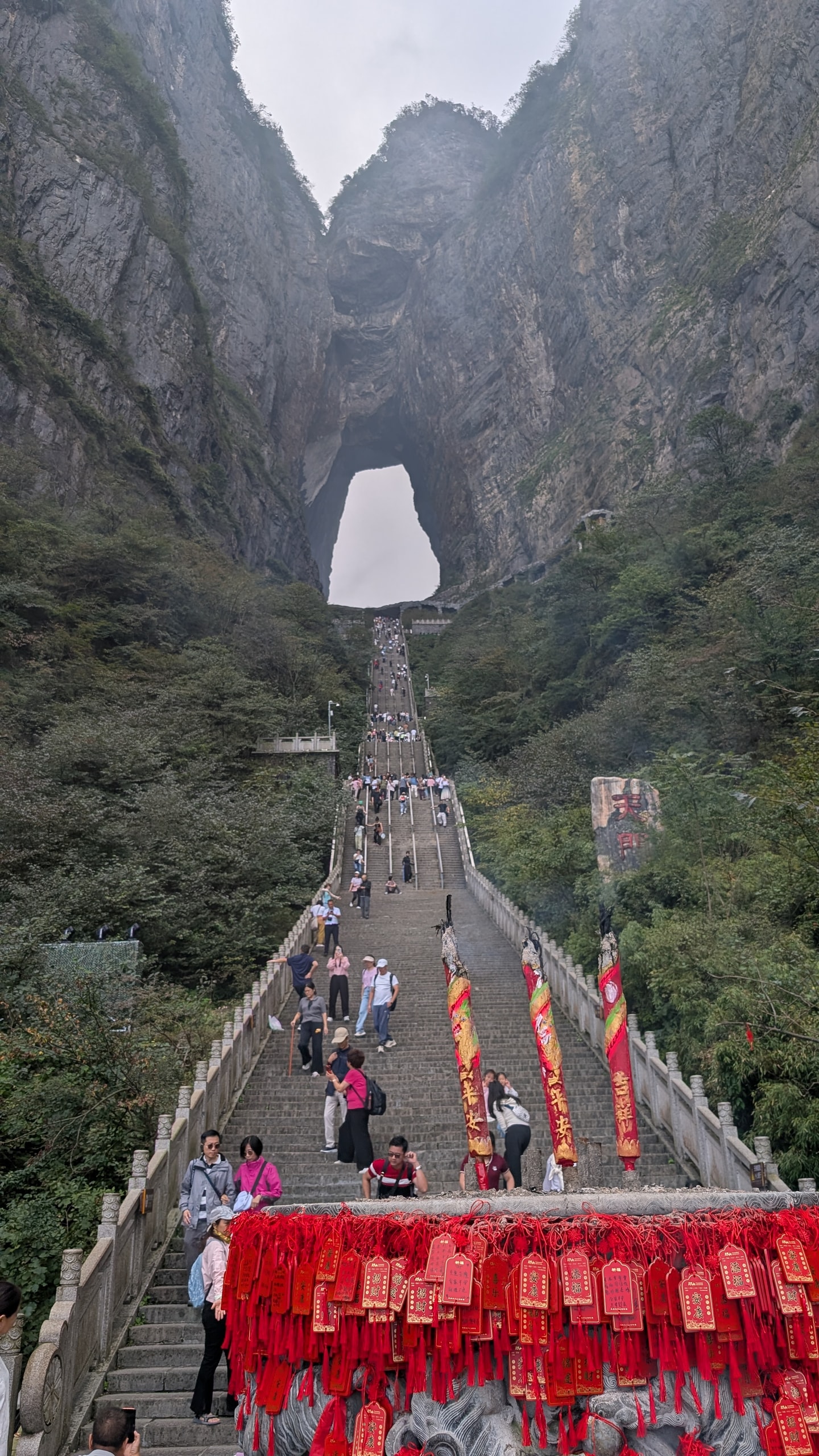 Red wooden wish tablets tied to a stone balustrade in the foreground with the staircase and Tianmen Cave arch behind