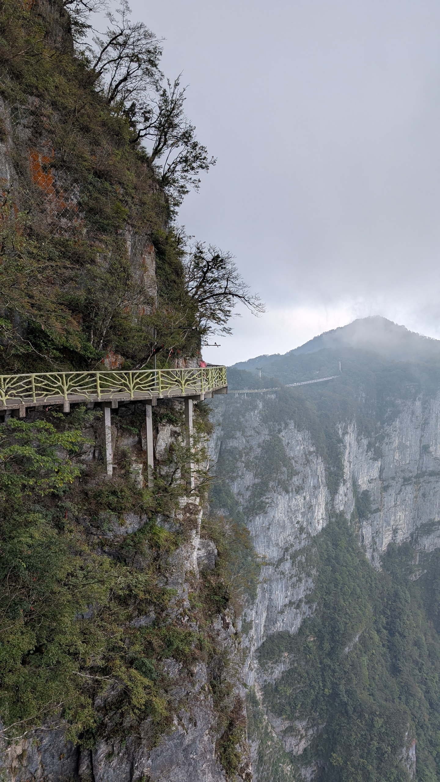 Timber walkway with yellow railings bolted to a vertical cliff face on Tianmen Mountain, and more walkway crossing toward a ridge in the distance