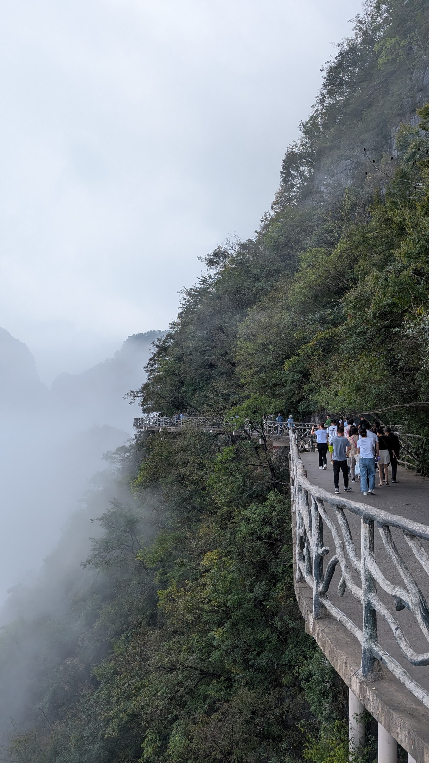 Visitors walking a narrow concrete cliff path with a fake-branch railing while fog fills the valley below on Tianmen Mountain