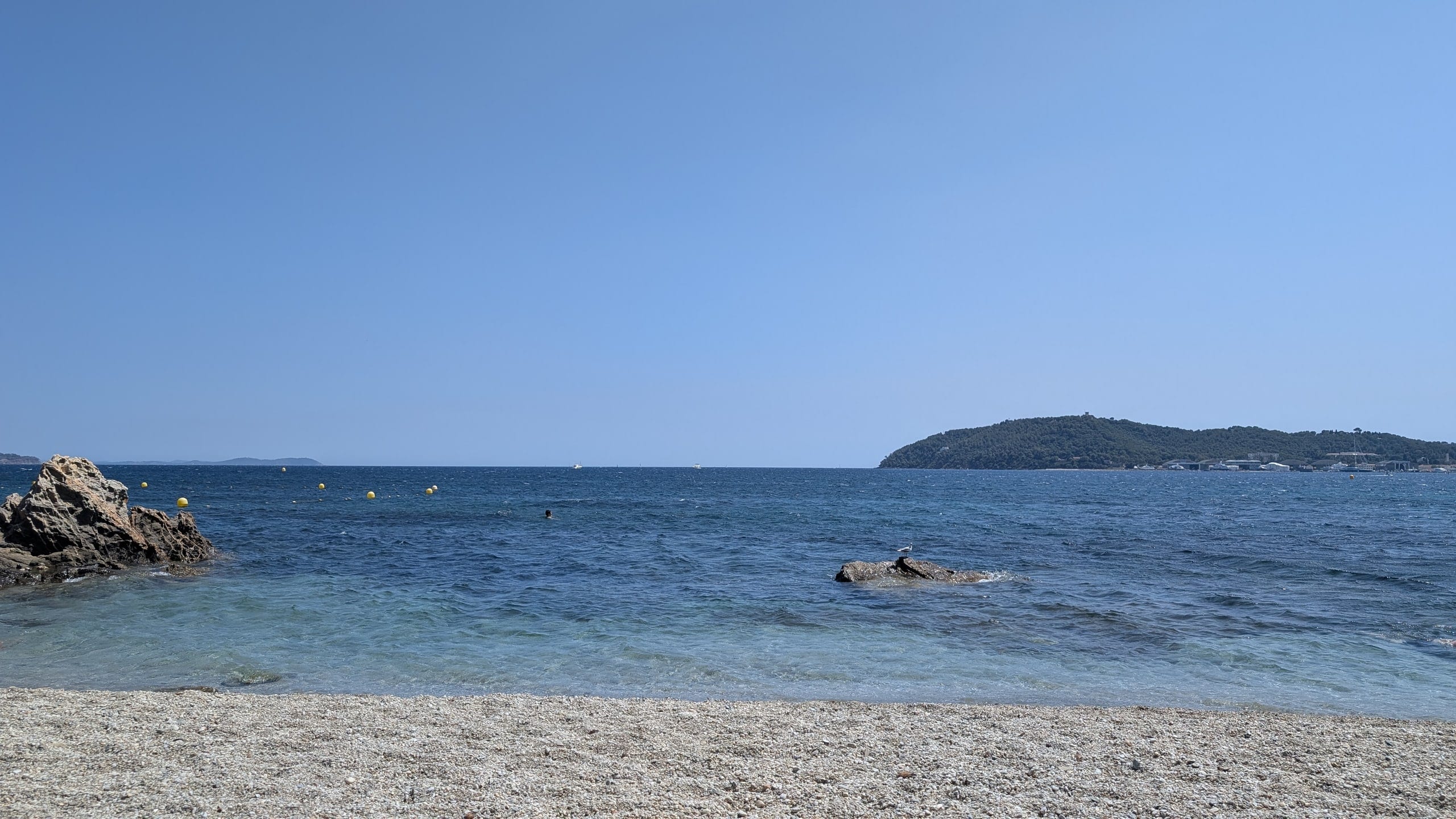 Clear shallow water over pebbles at a small beach on Toulon's eastern shore with a wooded headland across the bay