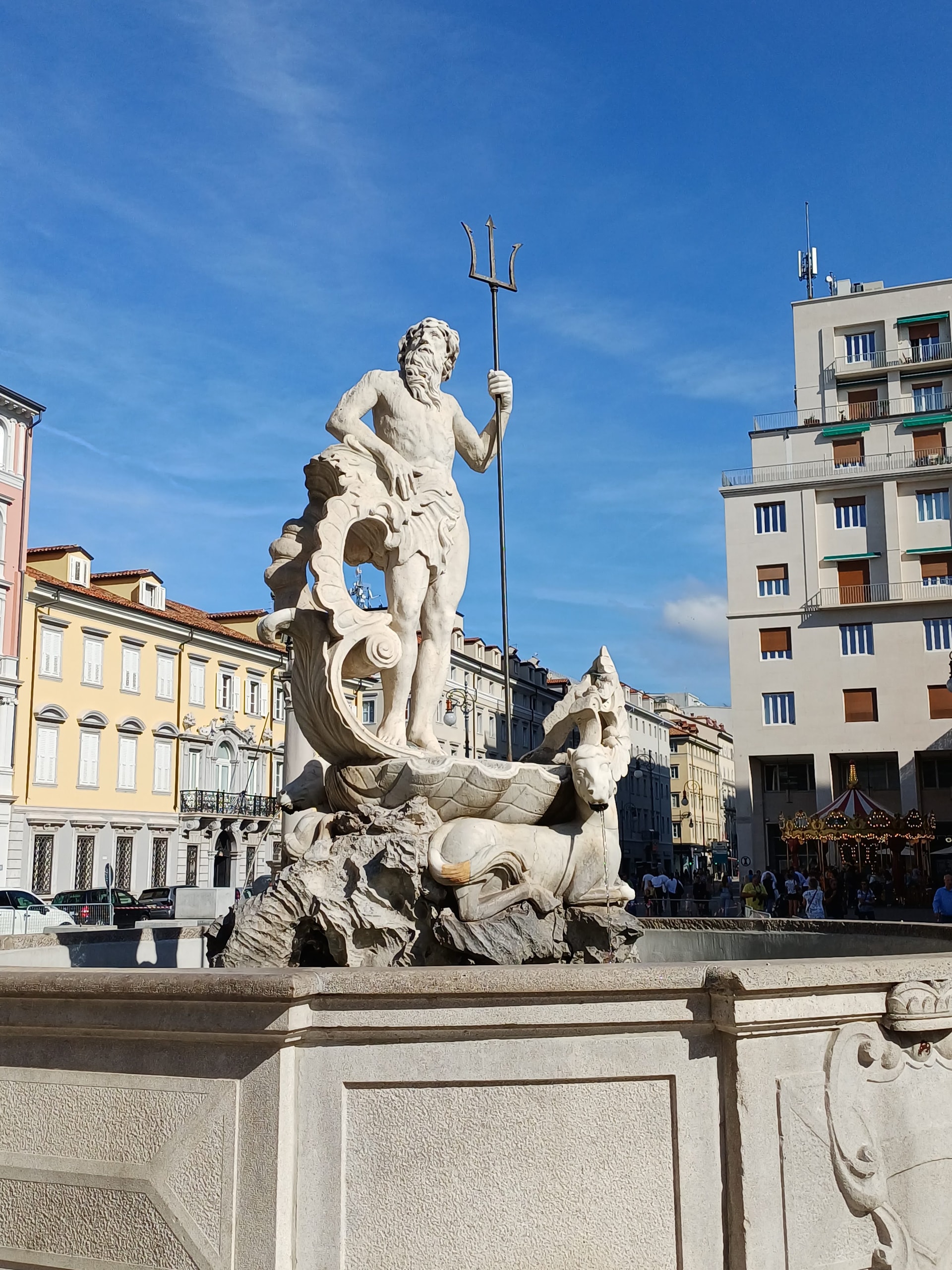 A white stone statue of Neptune holding a trident above a sea-horse fountain in Piazza della Borsa, Trieste, with a yellow palazzo and a carousel behind