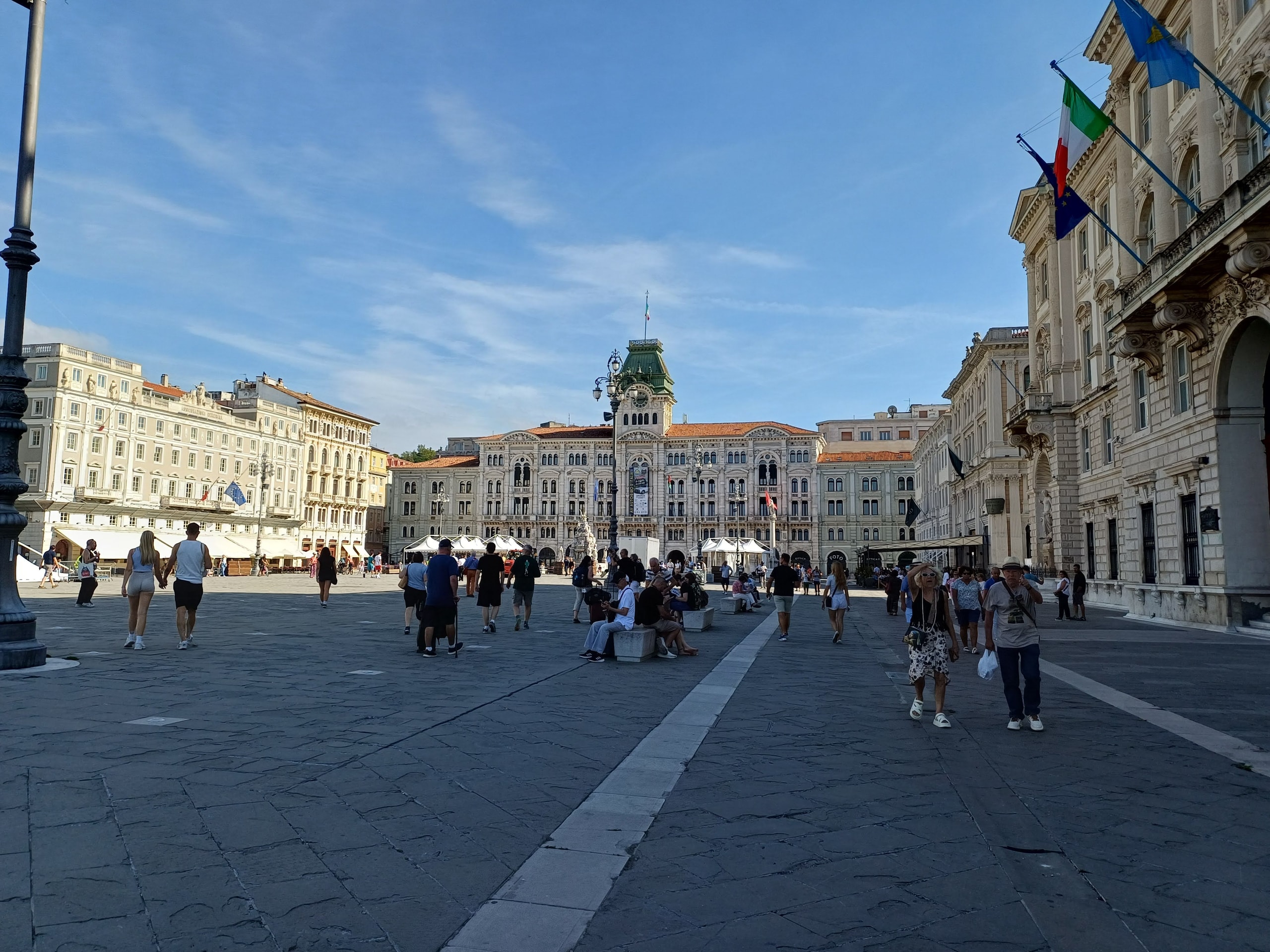 Piazza Unità d'Italia in Trieste in afternoon sun, with the town hall clock tower at the far end and Italian and EU flags on the government palace