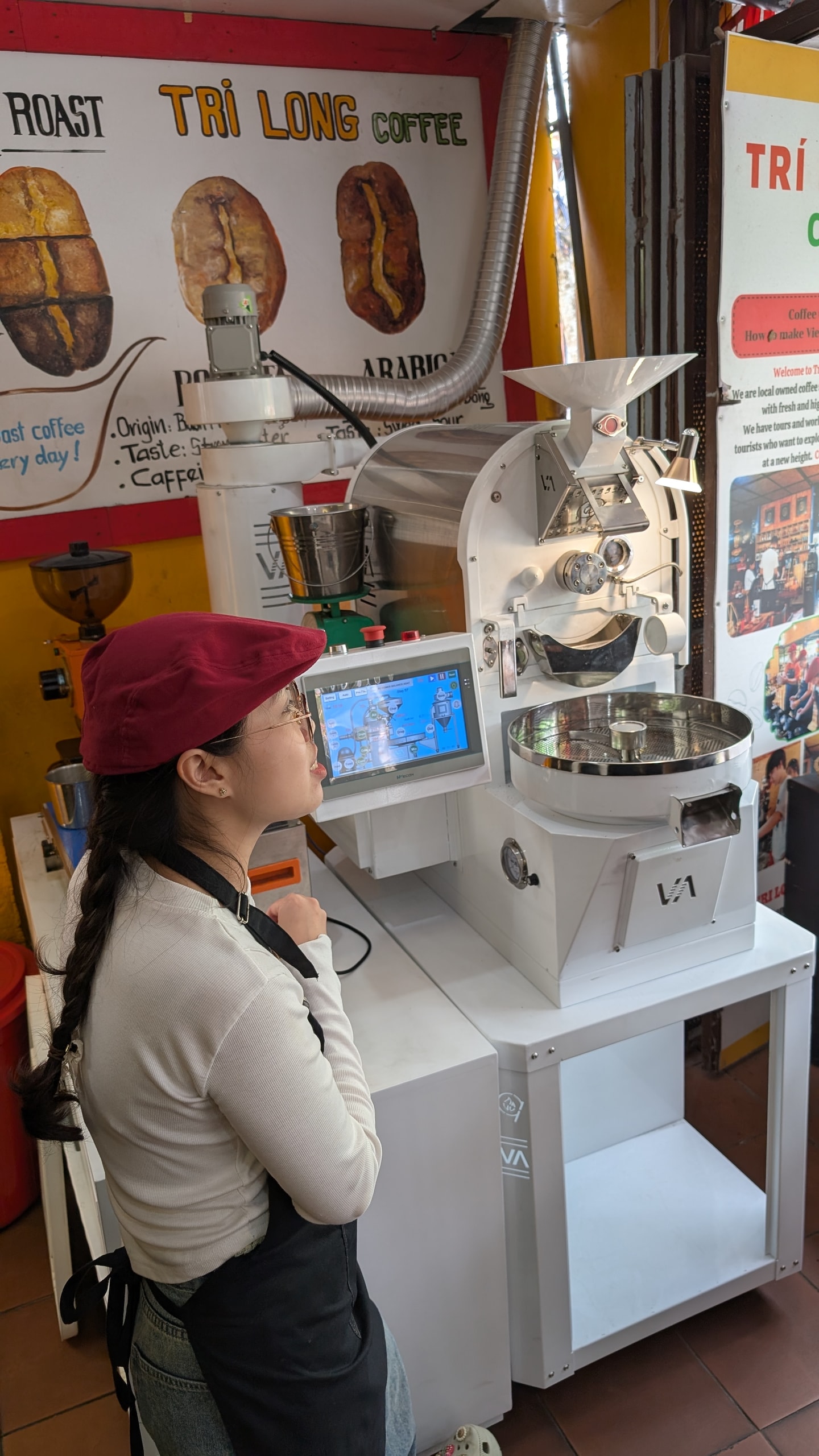A barista in a maroon cap watching a commercial coffee roaster's digital screen at Tri Long Coffee