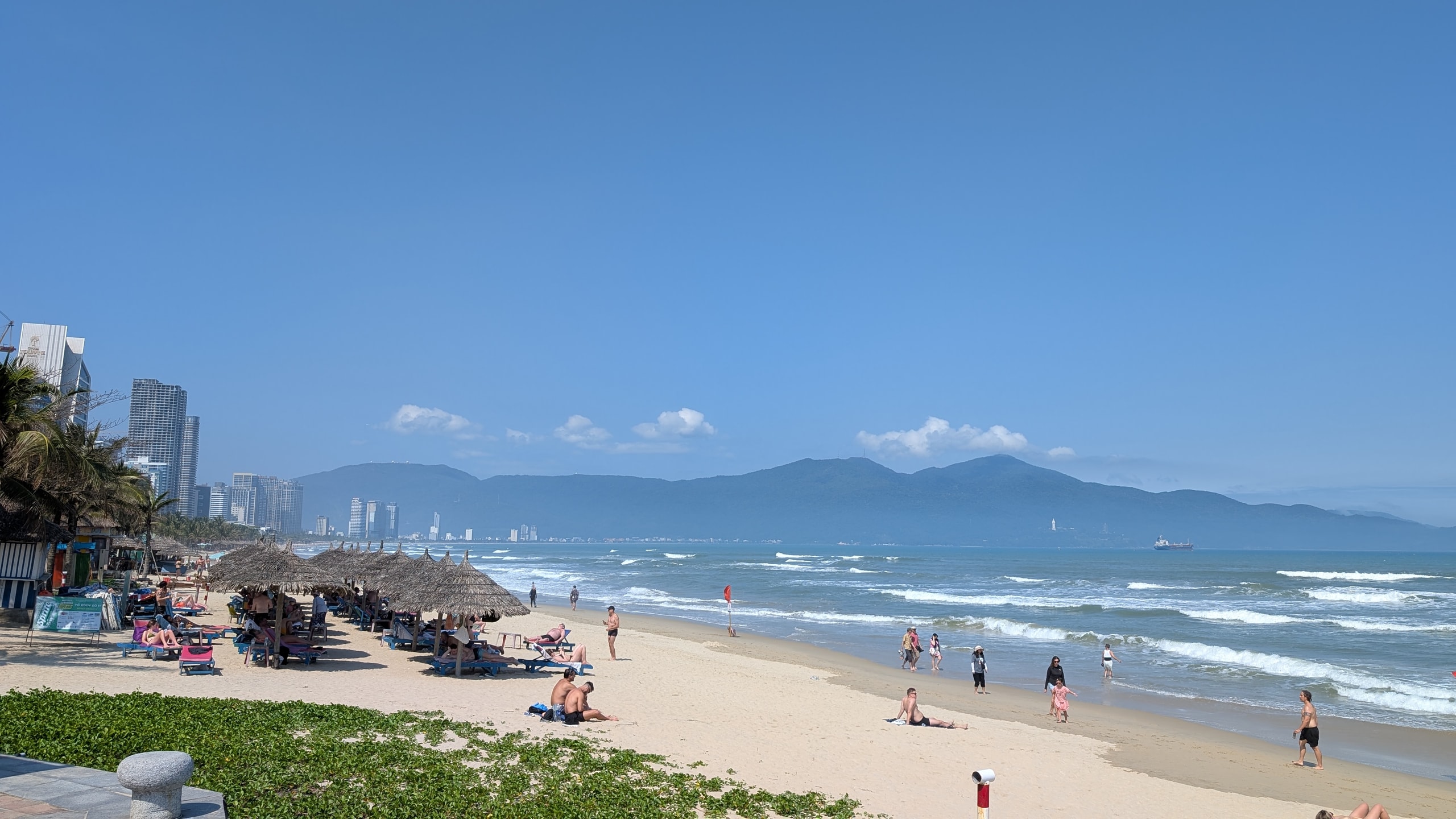 My Khe beach in Da Nang with thatched umbrellas, lounge chairs, blue water, and green mountains along the bay