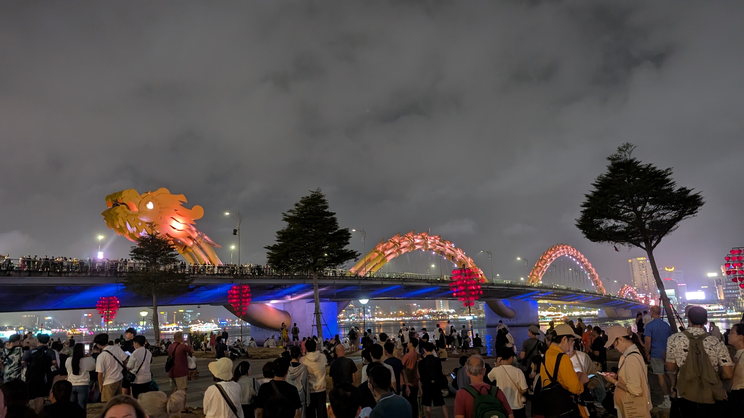 Crowd on the Da Nang riverfront watching the golden Dragon Bridge lit at night with blue underlighting