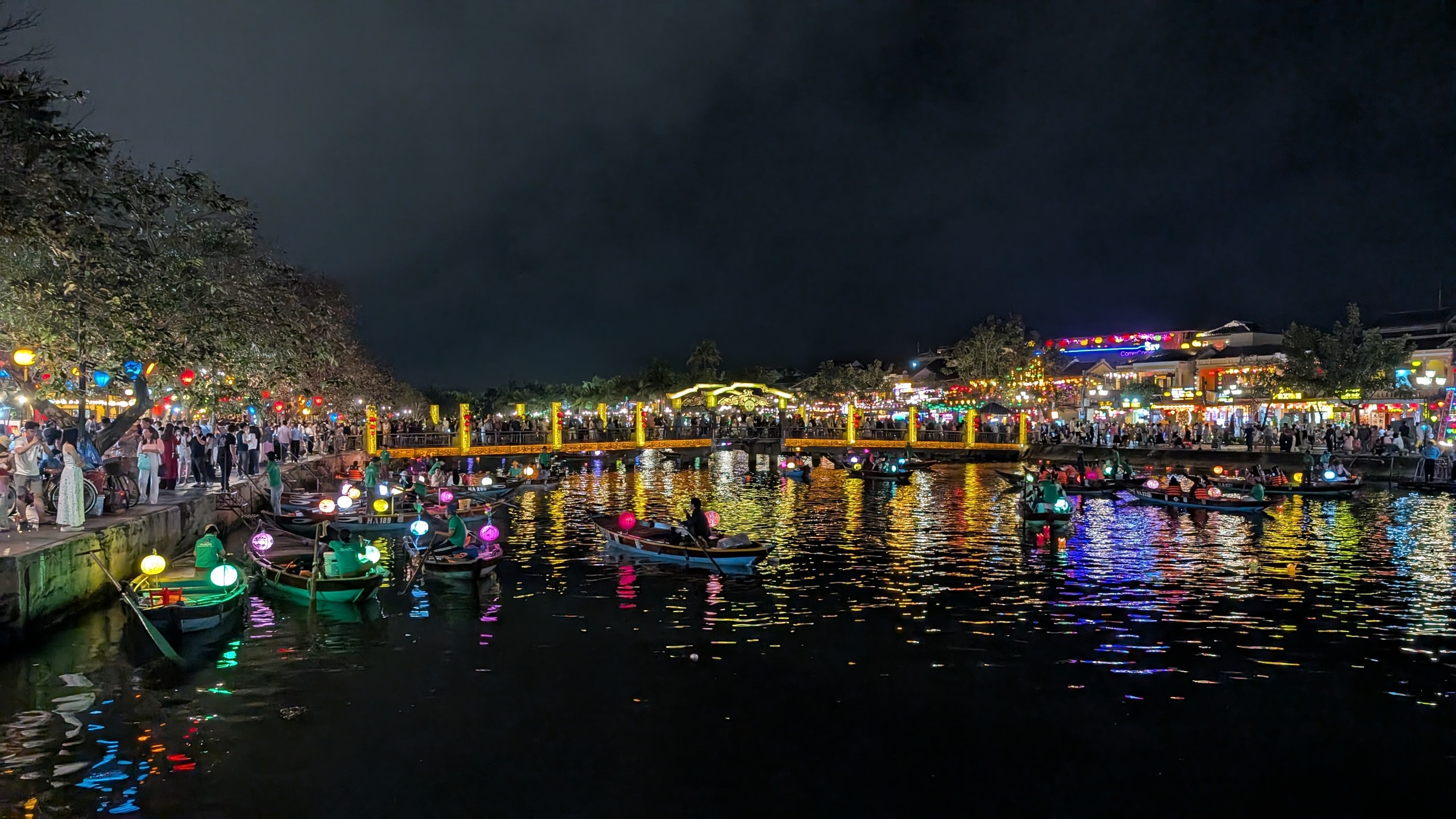 Lantern-lit boats on the Thu Bon River in Hoi An Ancient Town at night with a crowded bridge and neon-lit banks