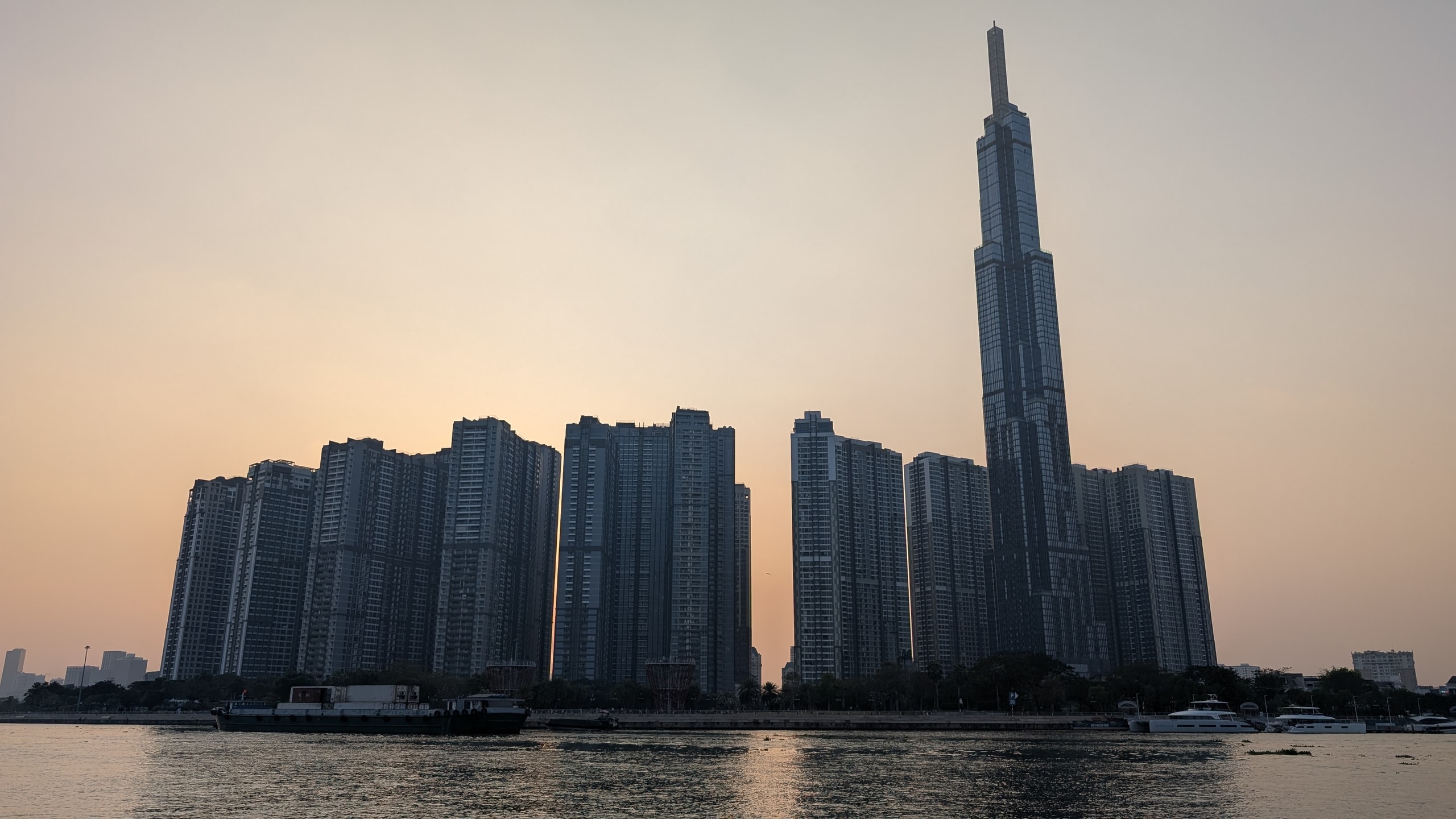 Landmark 81 and riverside towers silhouetted at dusk across the Saigon River with barges in the foreground