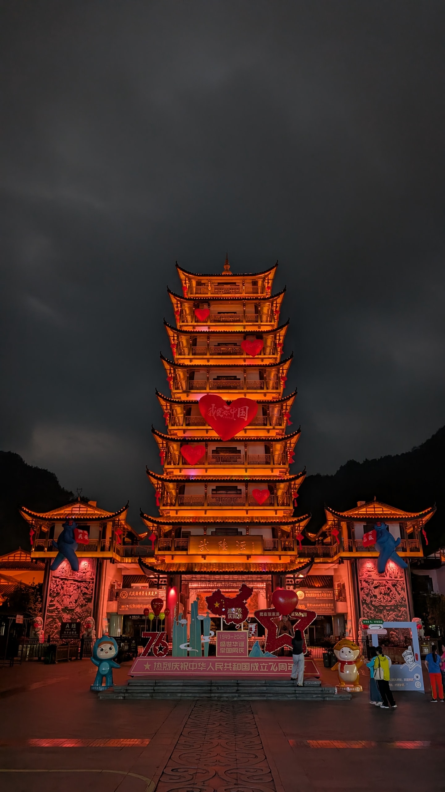 A tiered pagoda tower lit orange at night, hung with red heart decorations, on a plaza with a few people